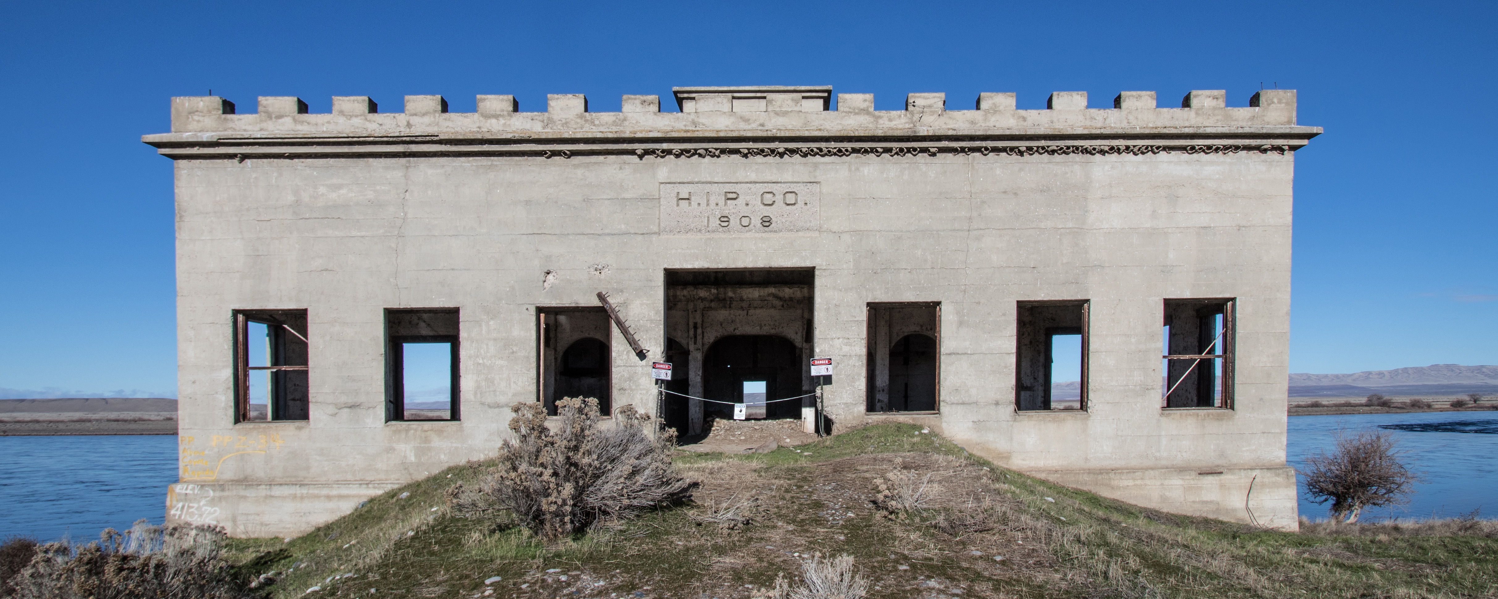 Symmetrical stone building with squared gaps at top like a castle wall, blue river and sky behind, with lettering “H.I.P. CO. 1908” above central doorway.
