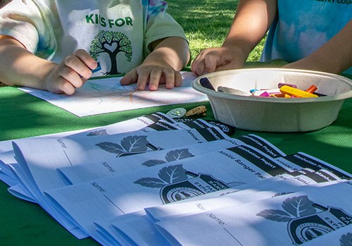 Kids sit at a table covered with a green tablecloth where the hands of two children coloring with crayons are visible.