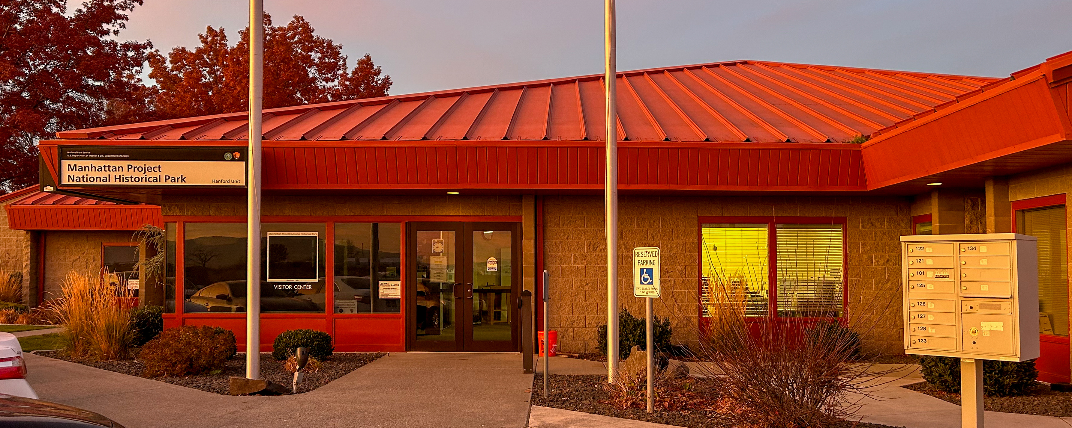 Single-level stone building with thick red-orange roof and window trim, double glass doors, paved sidewalk out front, sign with text, “Manhattan Project National Historical Park.”