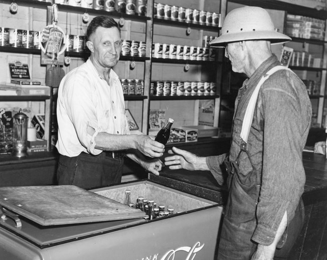A man hands a bottle to another man in a Wheat store.