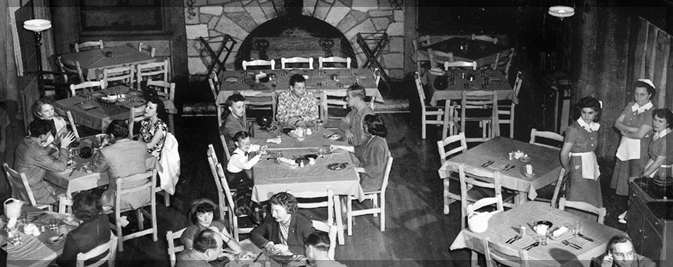 A black and white photo of people eating at several square tables. There is a stone fireplace in the background. Three women in aprons are standing nearby.