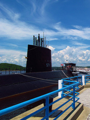 The Nautilus submarine next to a concrete dock. Blue sky and clouds in the background and blue railings in the foreground.