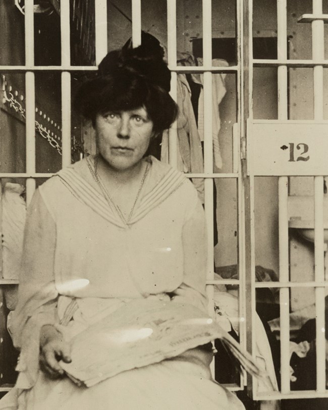 A woman looking tired and worn sits in front of prison bars