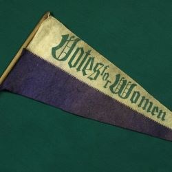 A purple and gold pendant with the words "Votes for Women" laying against a green background