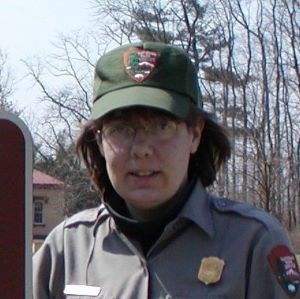 A park ranger in a hat smiling by a brown sign