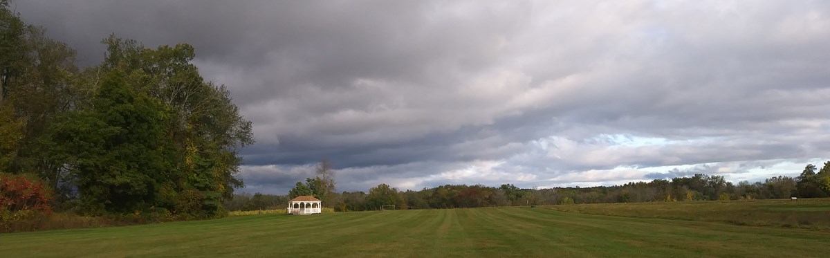 A photo of a grassy area with a gazebo, overlooking the farm with a cloudy sky