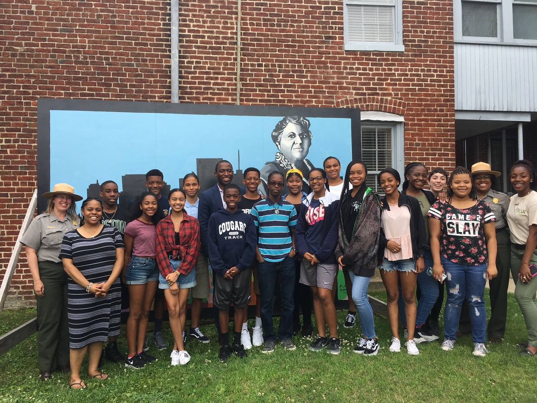 MLW Youth Summer Leadership Institute 2018 Students and Rangers pose in front of a mural of Maggie L. Walker in our courtyard
