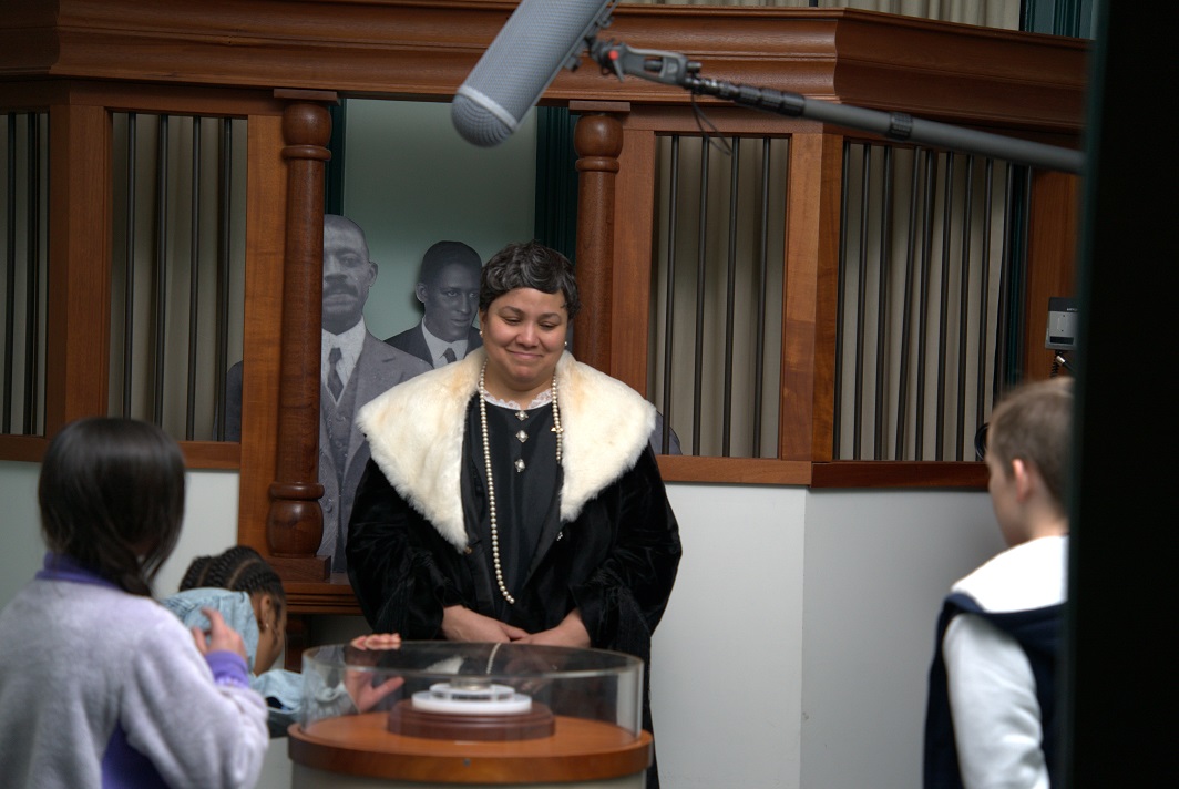 Maggie Walker Education Videos An actress in historical clothing of the early 20th century stands beneath a boom microphone
