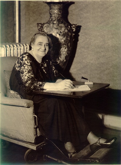 Mrs. Walker sitting in her rolling chair with an attached desk, with a paper notepad on the desk and a pen in her hand