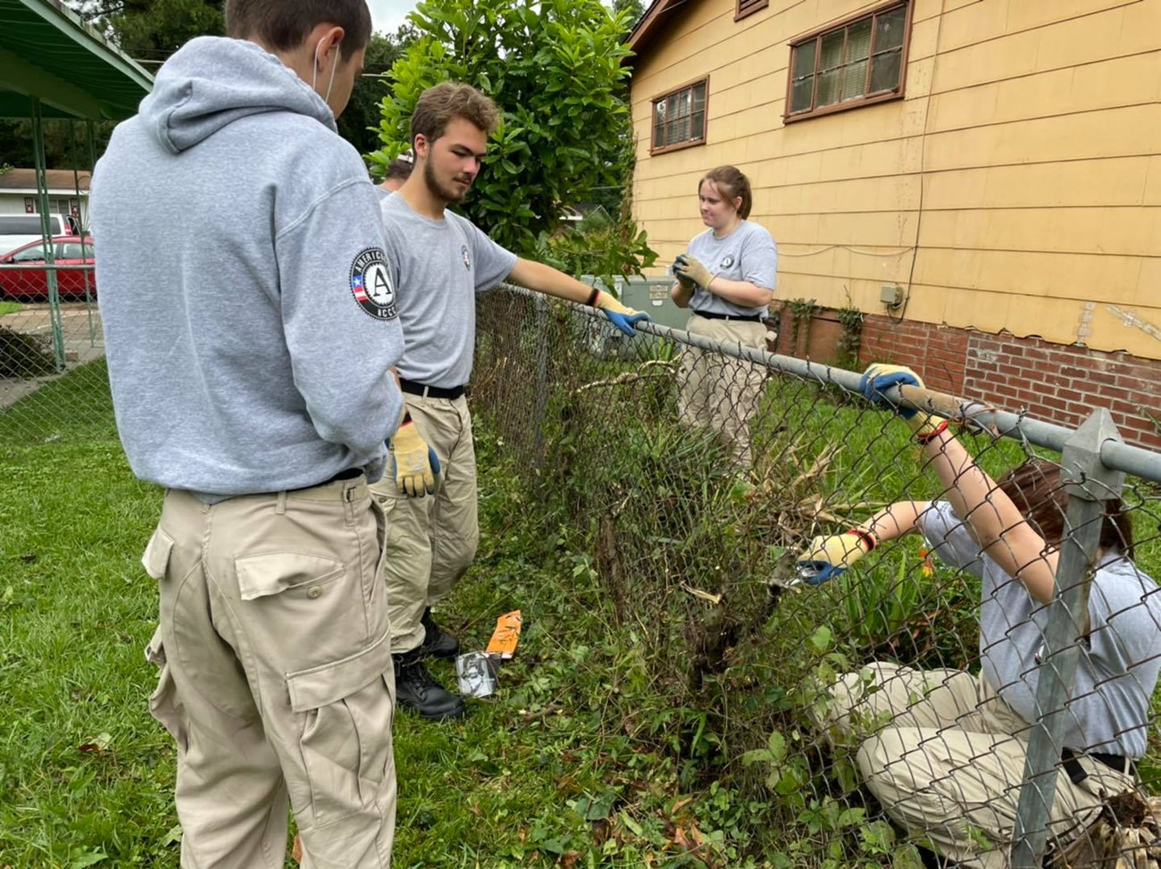 Four Americorps team members clear vegetation from chain link fence by Evers Home