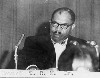 Mustachioed man in dark rimmed glasses speaks at hearing