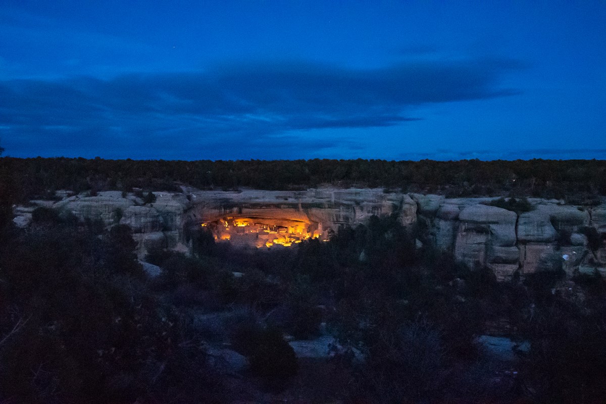 A twilight view into a sandstone canyon, with a stone-masonry cliff dwelling in a canyon lit up by lanterns