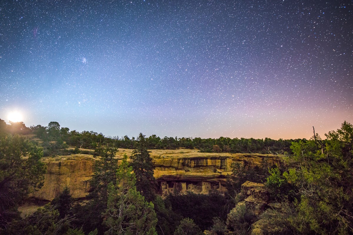 A full moon rises into a blue and starry sky over an ancient stone masonry village built into a cliff alcove in a forested canyon.