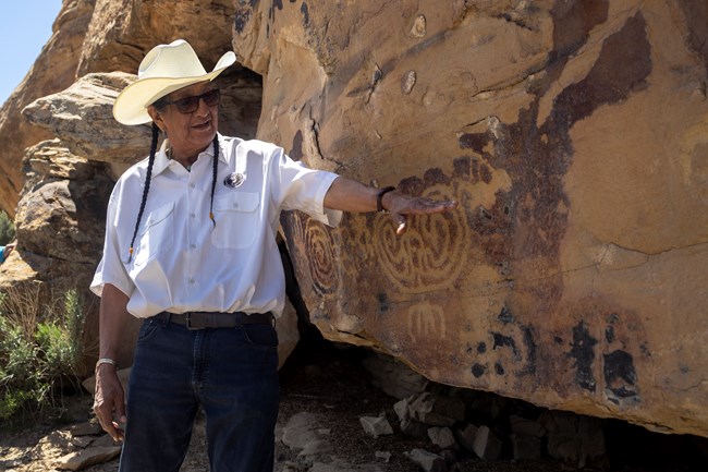 A man with a white cowboy hat, his hair in braids, talks as he gestures at a panel of petroglyphs (rock carvings) on a cliffside.