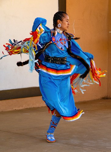 A Jicarilla Apache woman in a blue-orange dress dances with a fringed shawl billowing behind her.
