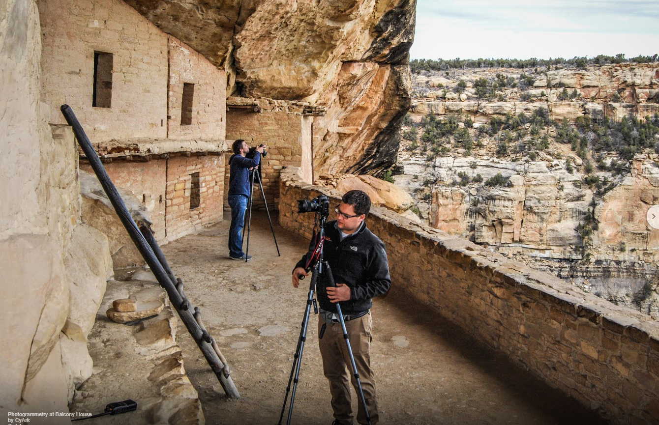 Two men work with cameras on tripods in a courtyard within an ancient stone masonry cliff dwelling village