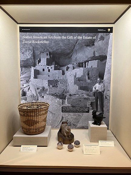 An exhibit with a 1900's Hopi wicker basket, a squirrel carving, three minature pots from mid-1900's and a blackware bird wihc is all black and shiny. This sit on a platform in front of a black and white photo of John D Rockefeller standing in a cliff d