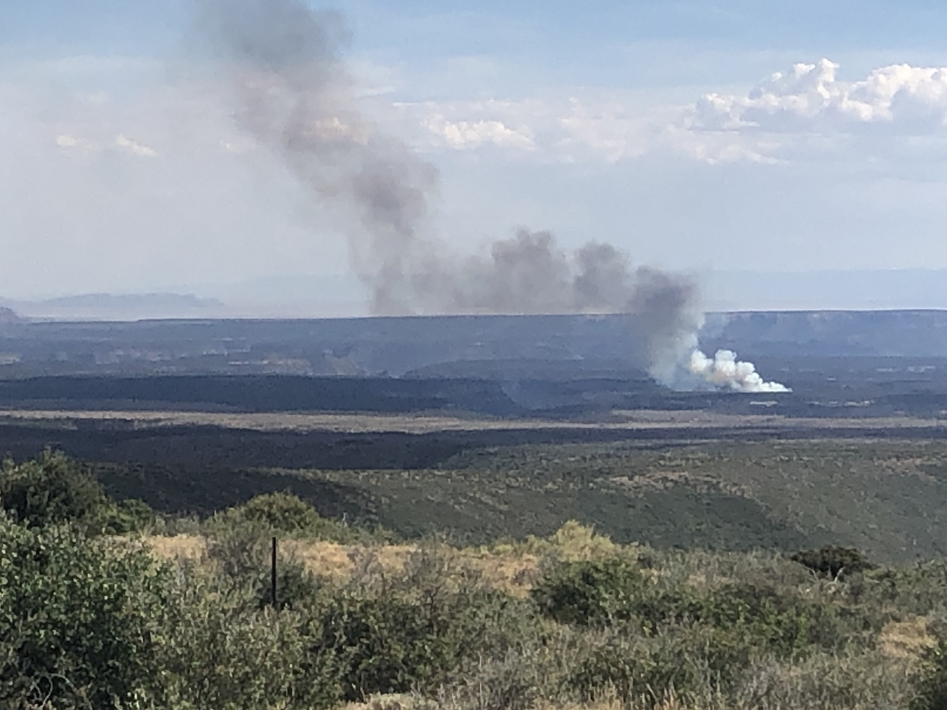 A plume of smoke rises from a canyon head, looking over the tilting landscape of Mesa Verde