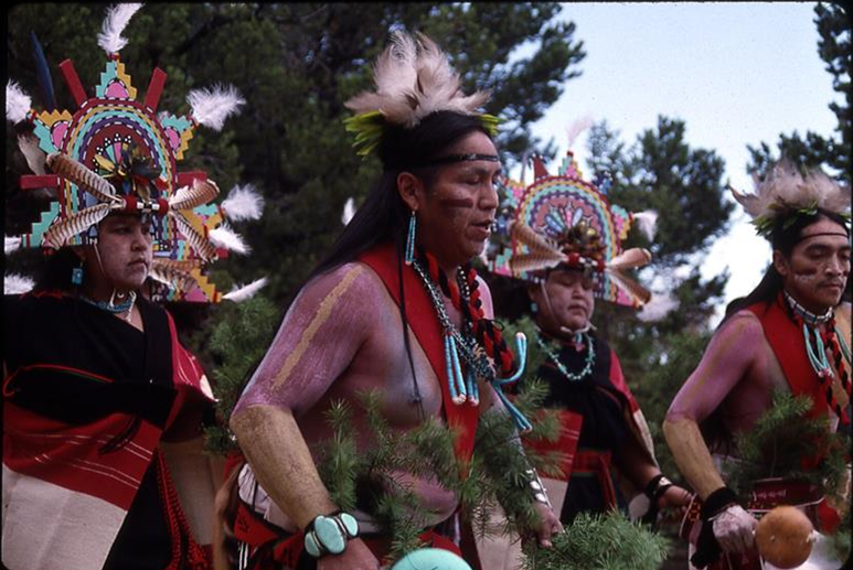 Four dancers in regalia dance dance with pinyon pines in the background
