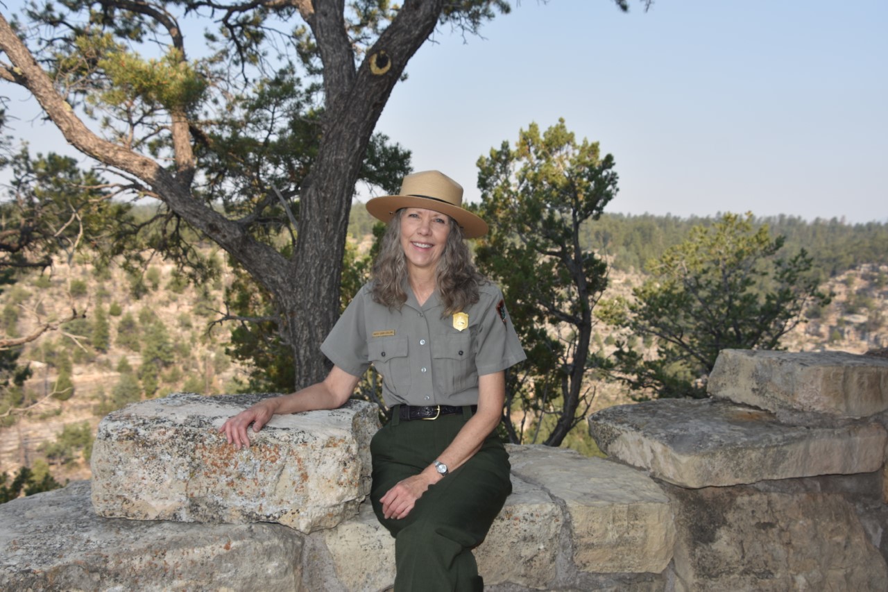 A woman in a park ranger uniform sits on a stone wall in front of a canyon