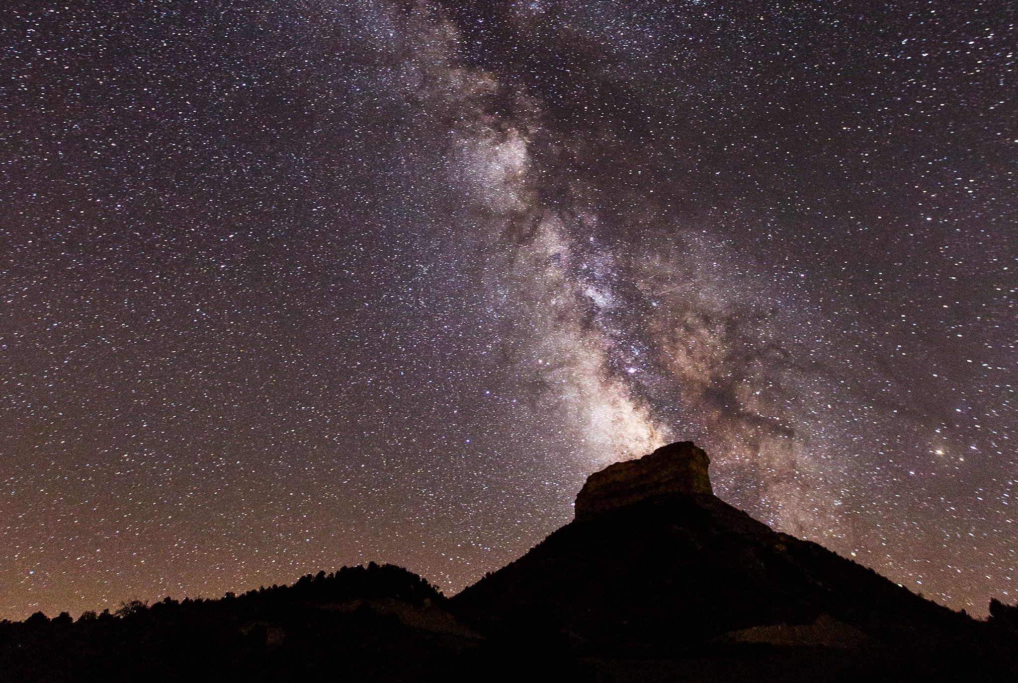 A nighttime view of a dark mesa and forested hills at the bottom of the frame. The sky above is full of thousands of stars. The glowing band of the Milky Way Galaxy seems to rise out of the mesa, spanning to the top left corner of the image.