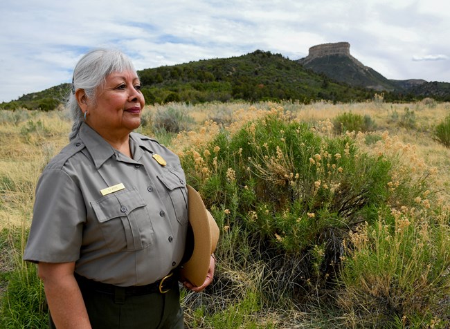A park ranger in her uniform with her hat under her arm stands in front of a chamisa bush, hills, and a prominent butte