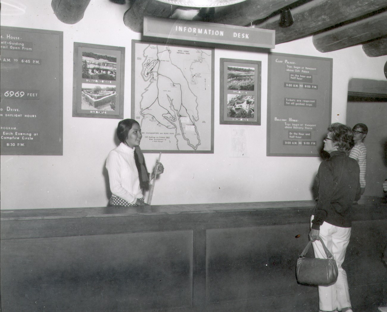 A young woman smiles at a park visitor from behind the museum information desk with maps and tour information posted on the wall