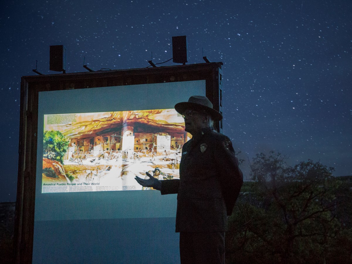 A park ranger stands gesturing in front of a projected screen outside with a starry night sky behind him