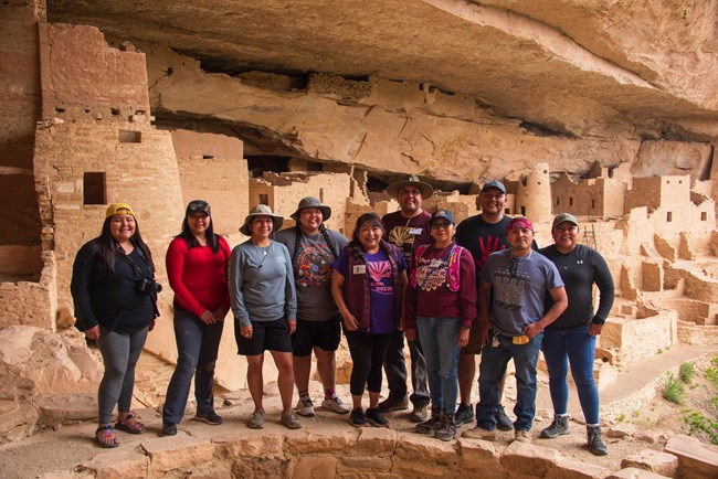 A group of ten Hopi visitors pose smiling in front of an ancestral stone masonry village