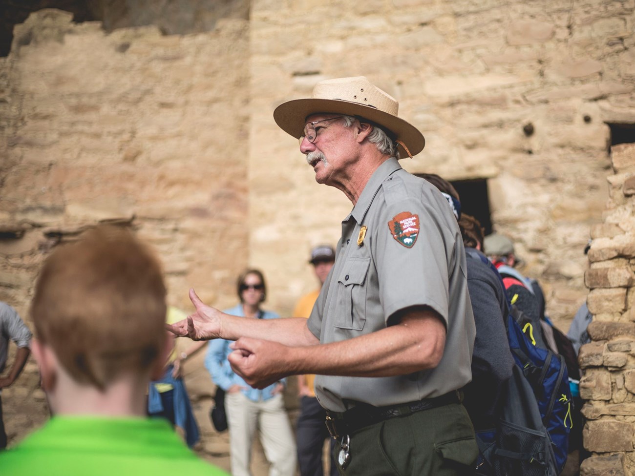 A ranger with white hair and mustache gestures with hands while visitors gather around listening