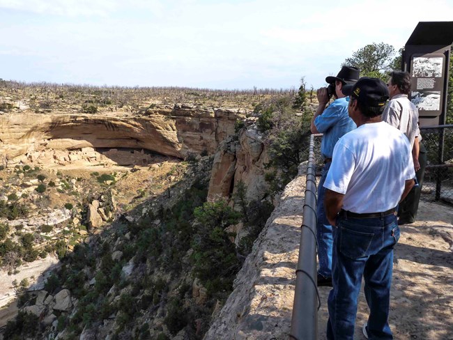 Three men stand at a railing at the edge of a canyon, looking across to a large ancient stone masonry village built into a sandstone alcove.