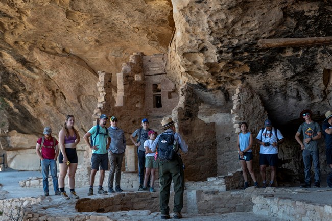 A rangers stands, talking with a group of ten visitors around at Balcony House, surrounded by stone structures and other pieces of Ancestral Pueblo history.