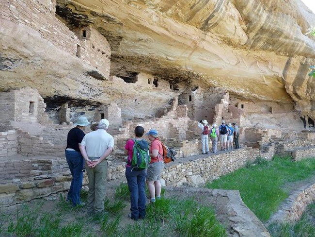 A group of visitors and a park ranger stand along the front of an ancient stone masonry village built inside a sandstone cliff.
