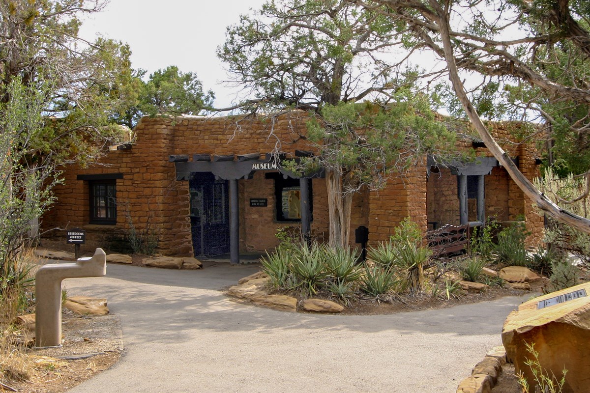 A paved path leads to a stone masonry museum building surrounded by yuccas and pinyon and juniper trees