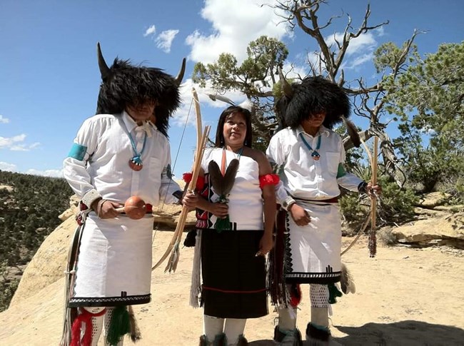 Three Pueblo dancers dressed in regalia, including bison headdresses, bows and arrows, and shell necklaces, stand on a sandstone outcrop with a canyon behind them