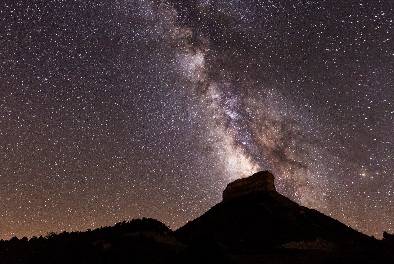 A starry sky bisected by the Milky Way rises over forested hills and the prominent mesa of Point Lookout