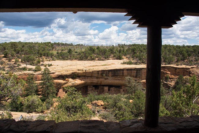 A view of a forested canyon and an ancient stone masonry village is seen through the opening of a porch with stone walls and carved wood column