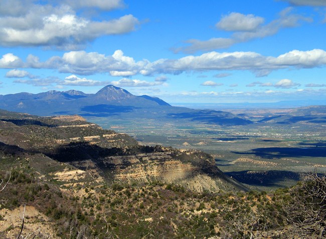 An expansive view from high elevation that exposes a partial mesa, the Ute Mountain Range, with the city of Cortez between and on a semi-cloudy day