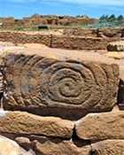 Spiral carved into a sandstone block in foreground with large, stone-masonry rooms in the background.