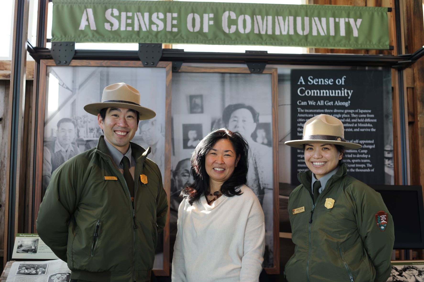 Three smiling Japanese American people stand inside the Minidoka Visitor Center in front of an exhibit. The middle-aged woman in the middle wears a white sweater, and the young man and young woman alongside her wear park ranger hats and uniforms.