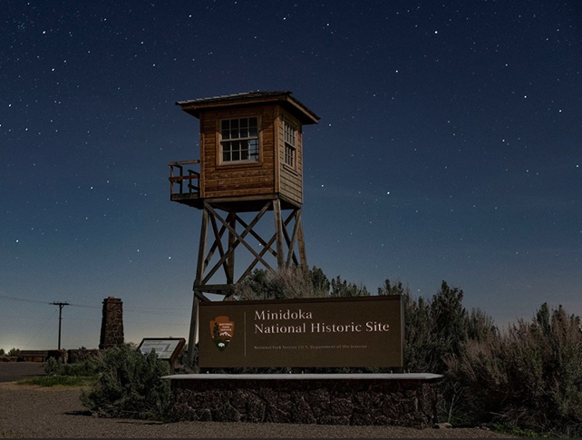 MIIN-Tower-Stars_Stan-Honda Behind the Minidoka National Historic Site park entrance sign, a tall wooden guard tower stands in front of a starry sky.
