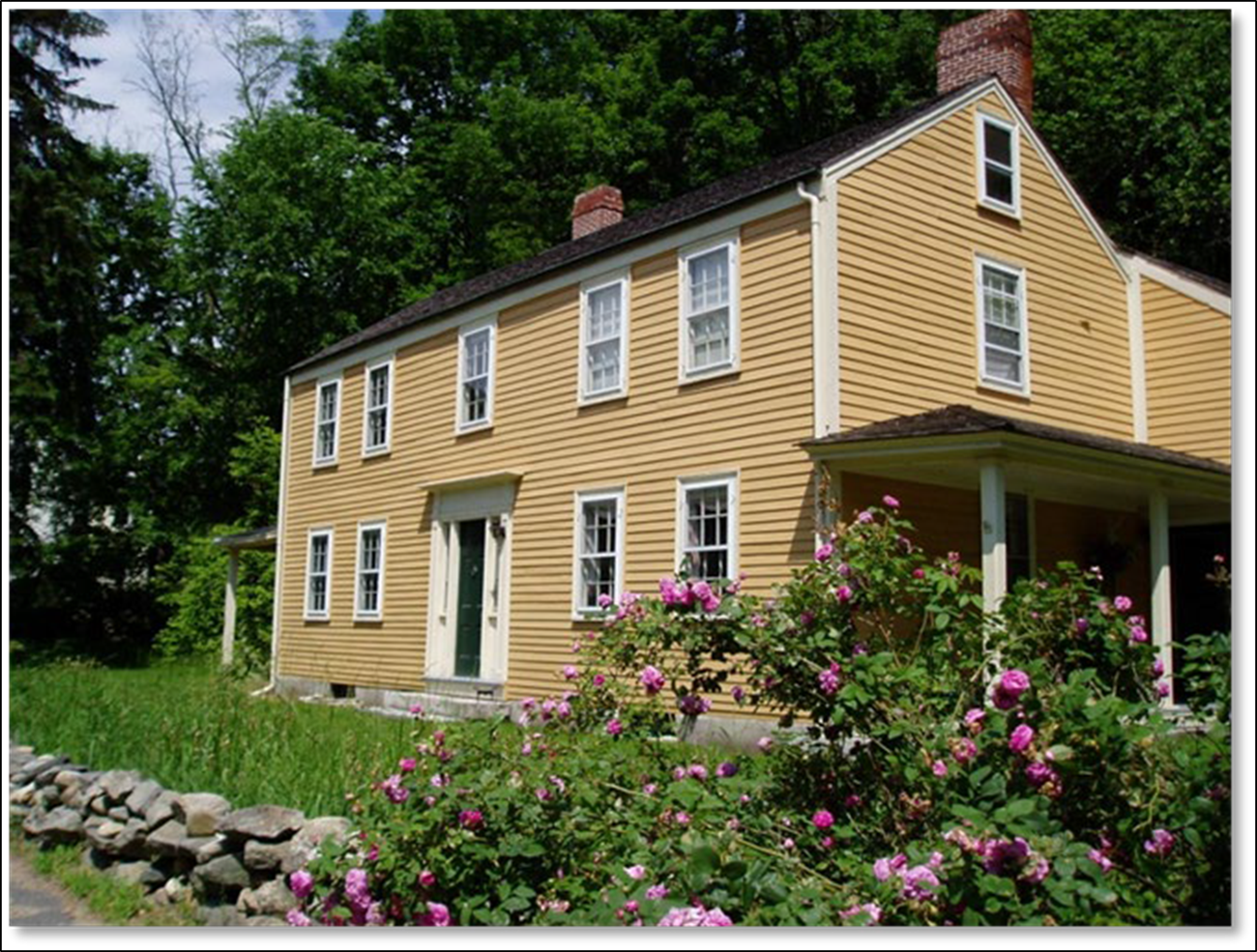 A yellow colonial era house made of wood with two stories and a central door. Surrounded by green trees and a stone wall.