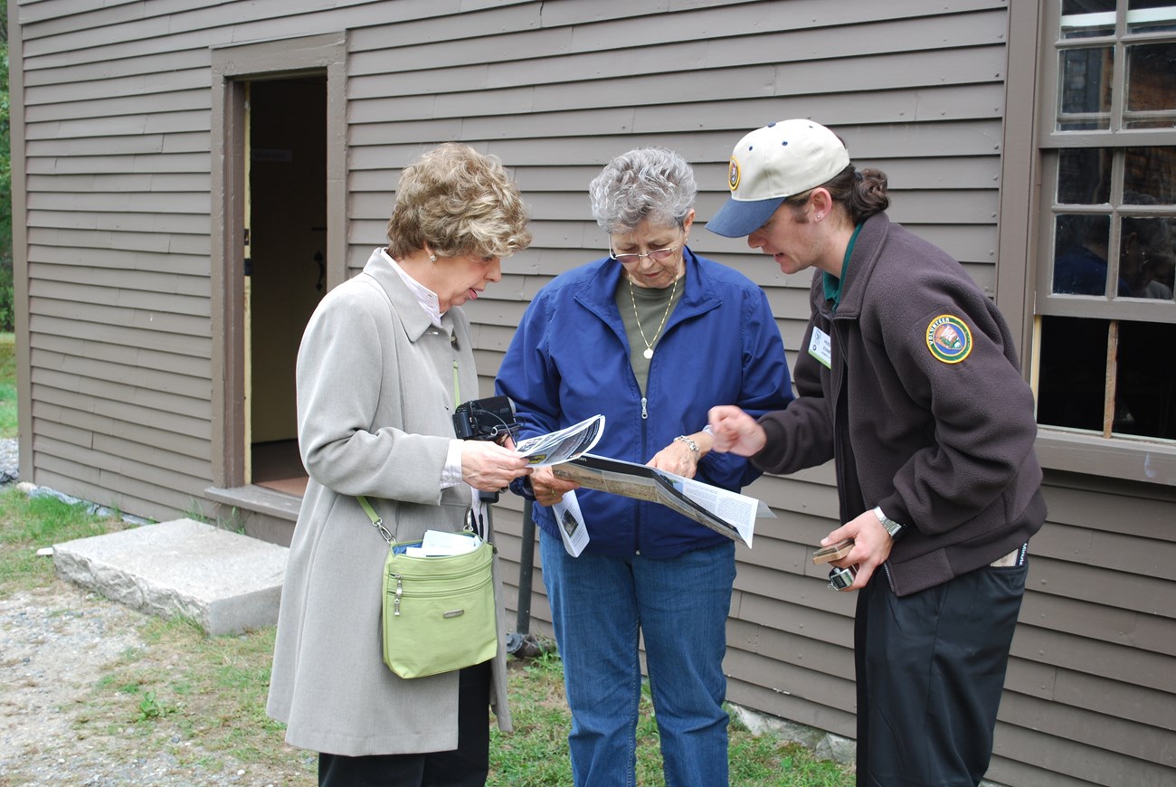A volunteer in brown sweater gives directions to two visitors holding a map. The group stands in front of a wooden historical house.