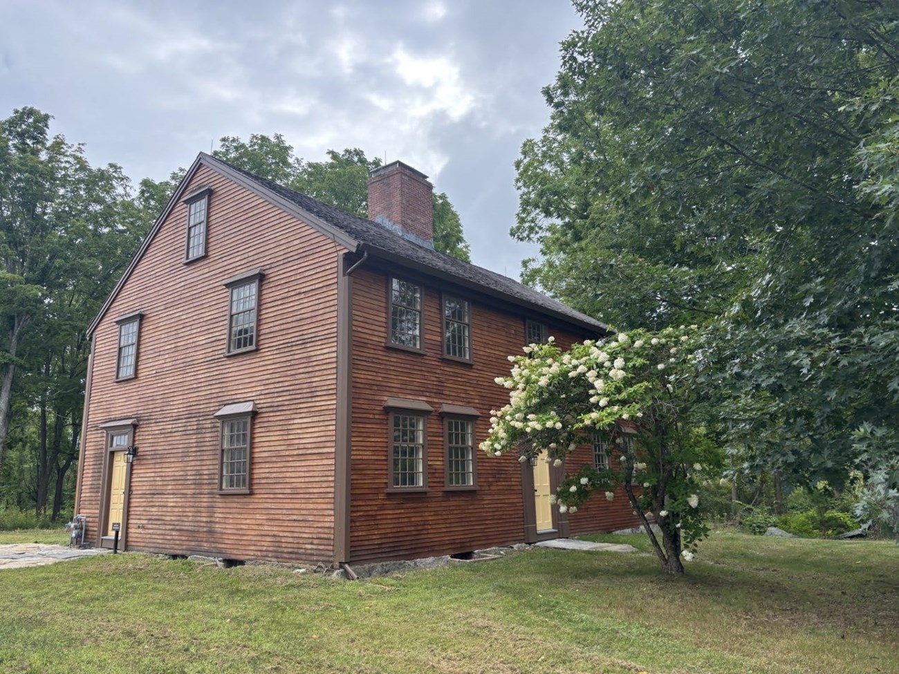 A wooden 1786 house with central chimney. Surrounded by trees and green grass.