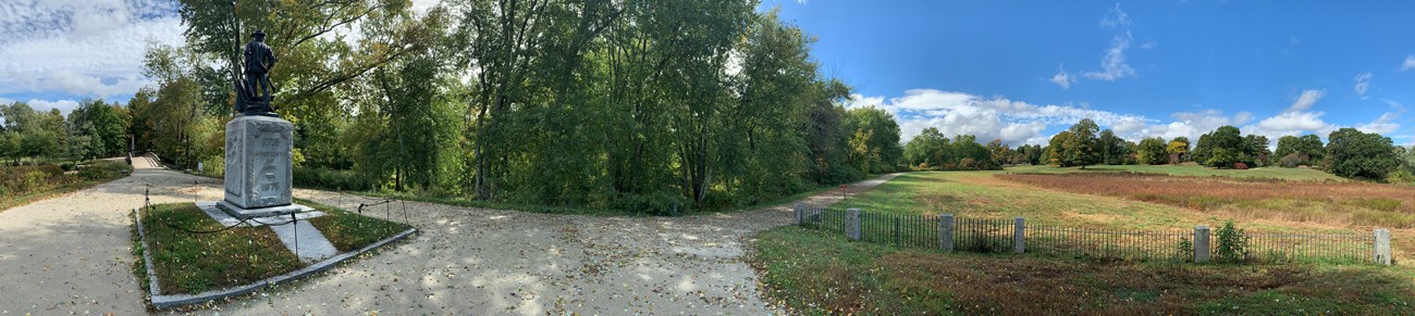 A wooden bridge crosses a river at left. A dirt trail extended right from the bridge to a stone and metal monument of a Minute Man. The dirt trail continues right, running along a green tree line through a large open field and bends up onto a hillside.