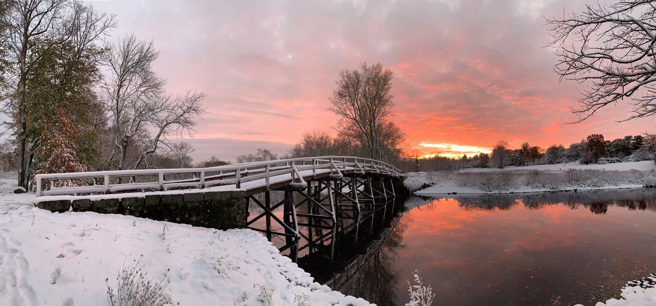 A long wooden bridge is covered in a fresh layer of white snow. The scene is illuminated by a fiery orange sunset that reflects off of the snow and water under the bridge. There are trees at either end of the bridge with no leaves.