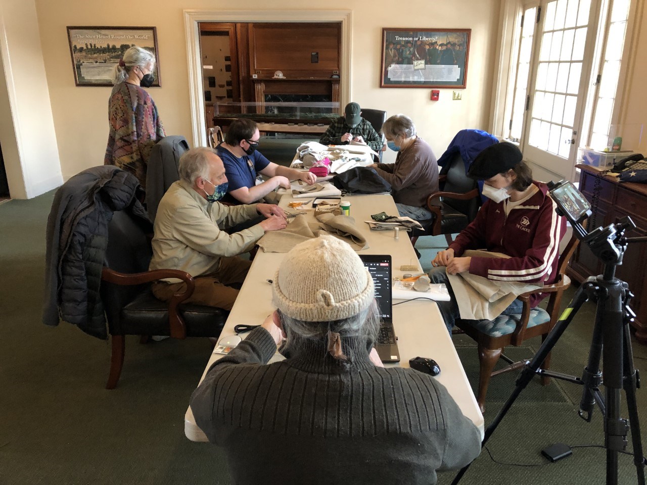 Several people sit at a long table sewing in a sunlit room