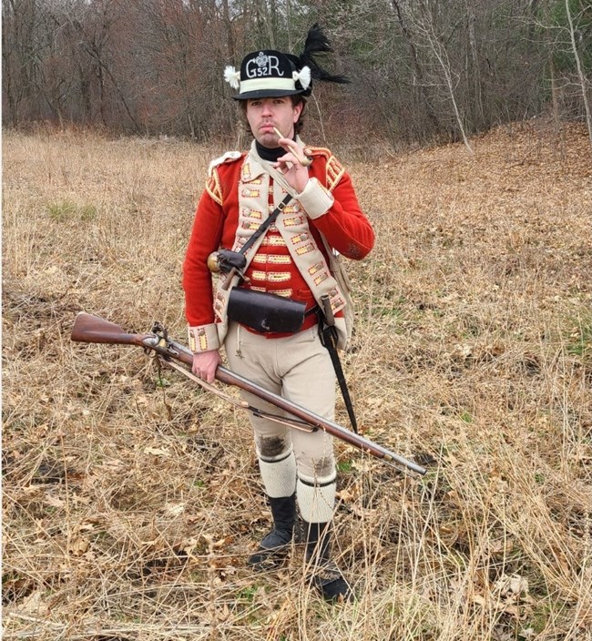 A Revolutionary War British soldier in a short red coat with a black cap and musket stands in a field smoking a pipe
