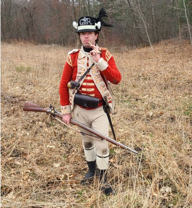 Light Infantry soldier 52nd Foot A Revolutionary War British soldier in a short red coat with a black cap and musket stands in a field smoking a pipe