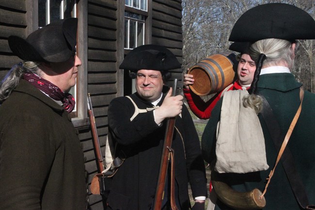 Four men dressed in colonial clothing and armed with muskets, one holding a small barrel standing outside a wooden colonial house